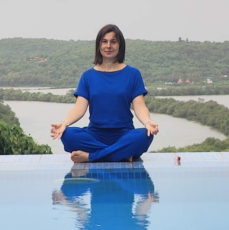 Yoga therapist Emilie sitting in easy pose (Sukhasana) on an infinity pool edge overlooking a river landscape, India